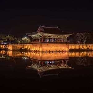 Un pavillon traditionnel et un mur de pierre, illuminés la nuit, projetant un reflet parfait sur l'eau calme en contrebas.