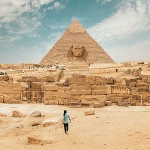 Une personne marche vers le Grand Sphinx et une grande pyramide à travers une étendue sablonneuse de ruines antiques sous un ciel partiellement nuageux.