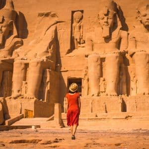 Eine Frau in einem roten Kleid und Strohhut geht auf einen großen, antiken Tempel zu, der in eine Klippe mit kolossalen sitzenden Statuen gemeißelt ist.