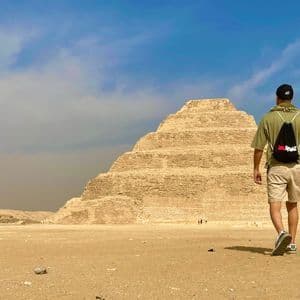 Ein Mann mit einem WeRoad-Rucksack wandert durch eine Wüste auf eine große Stufenpyramide zu.