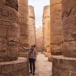 Un homme se tient dans une salle de temple antique, levant les yeux vers les hiéroglyphes gravés dans les imposantes colonnes de pierre.