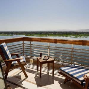 Ein sonniger Balkon mit gestreiften Loungemöbeln und einem kleinen Tisch, mit Blick auf einen breiten Fluss und ferne grüne Ufer.