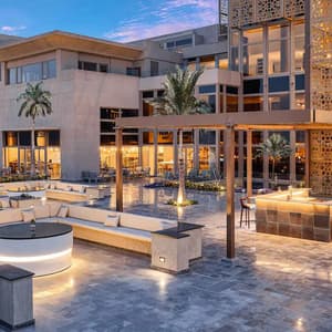 An outdoor terrace of a modern hotel at dusk, featuring illuminated seating areas, a bar, and palm trees.