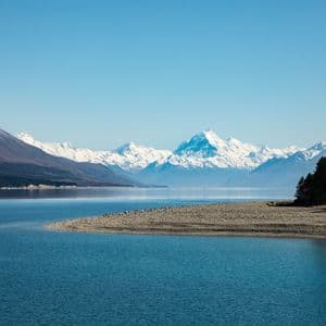 Un lac bleu et calme avec une plage de galets au premier plan, et une chaîne de montagnes enneigées en arrière-plan sous un ciel dégagé.