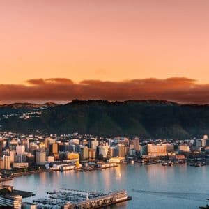 Vue plongeante sur une ville côtière et son port, au pied de collines verdoyantes, baignée par un chaud coucher de soleil orangé.