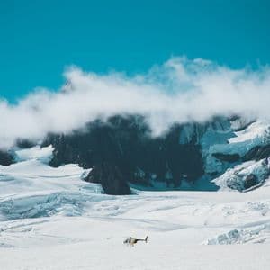 Un hélicoptère se repose sur un vaste glacier au pied de montagnes enneigées sous un ciel bleu.