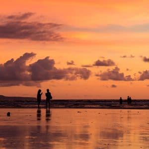Silhouettes d'un voyage en groupe WeRoad sur une plage mouillée au coucher du soleil, avec un ciel orange et des nuages se reflétant dans le sable.