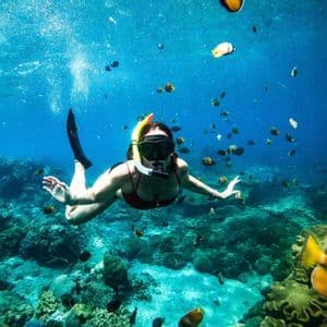 Une femme en maillot de bain rouge fait de la plongée avec tuba sous l'eau, nageant parmi un banc de poissons au-dessus d'un récif corallien.
