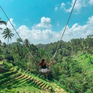 Une femme sur une grande balançoire contemple de luxuriantes rizières en terrasses et une forêt tropicale sous un ciel bleu.