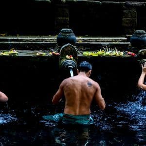 Un voyage de groupe WeRoad vu de dos participant à un rituel de purification sous des fontaines d'eau en pierre dans un bassin sombre.