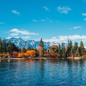 Une vue sur un lac calme montre un rivage avec des arbres d'automne colorés et des montagnes enneigées en arrière-plan.