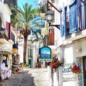 Una stretta strada acciottolata si snoda tra edifici imbiancati con una palma alta, negozi e balconi sotto un cielo azzurro brillante.