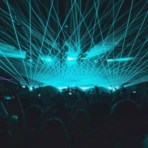 Eine Menge silhouettierter Menschen in einem dunklen Veranstaltungsort unter einer lebhaften, sich kreuzenden cyanfarbenen Laserlichtshow.