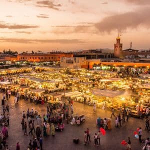 Une vue plongeante sur une place de marché animée au coucher du soleil, remplie de monde se promenant entre des étals illuminés.