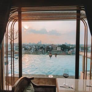 Dos personas en una piscina infinita en la azotea, con vistas a la ciudad al atardecer, observadas desde el interior de una habitación con una copa de vino sobre una mesa.