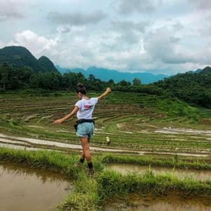A woman from a WeRoad group trip balances on a narrow, grassy path between flooded rice paddies with green mountains in the background.