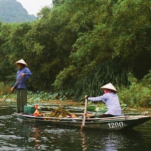 Dos personas con sombreros cónicos navegan en un bote por un río, rodeado de exuberante vegetación y colinas; uno impulsando con pértiga y el otro remando.