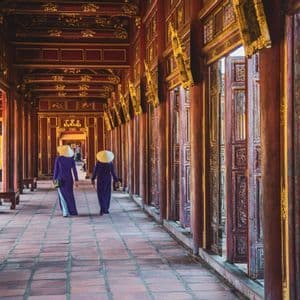 Deux femmes coiffées de chapeaux coniques et vêtues de robes traditionnelles violettes marchent le long d'un long couloir orné de piliers en bois rouges et dorés.