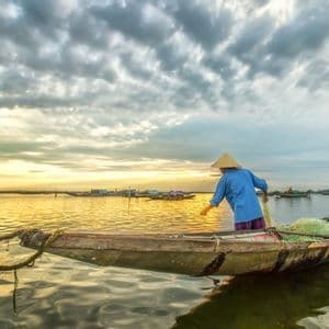 A fisherperson in a conical hat handles a fishing net on a small wooden boat at sunset under a cloudy sky.