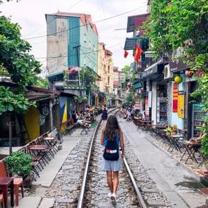 A woman with a WeRoad backpack walks along a train track that runs through a narrow city street lined with outdoor cafes.
