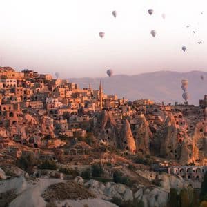Globos aerostáticos flotan sobre un pueblo construido en acantilados rocosos y casas cueva durante un amanecer brumoso.