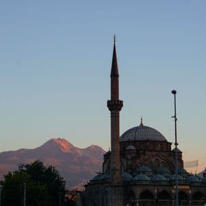 Una gran mezquita con dos minaretes y varias cúpulas se alza frente a una cordillera iluminada por el sol contra un cielo despejado.