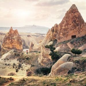 A lone person in an orange top stands with arms raised, overlooking a rocky valley with conical formations and cave dwellings.