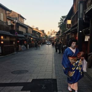 Une femme vêtue d'un kimono bleu traditionnel et maquillée de blanc marche le long d'une rue pavée bordée de bâtiments traditionnels en bois au crépuscule.