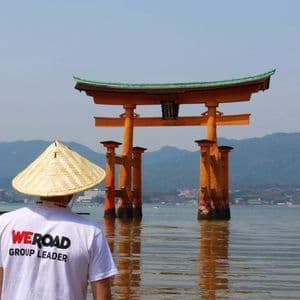 Ein WeRoad-Reiseleiter mit konischem Hut blickt auf ein großes orangefarbenes Torii-Tor, das im Wasser steht.