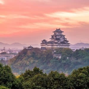 Eine traditionelle japanische Burg auf einem bewaldeten Hügel, die eine Stadt mit nebligen Bergen bei Sonnenuntergang überblickt.