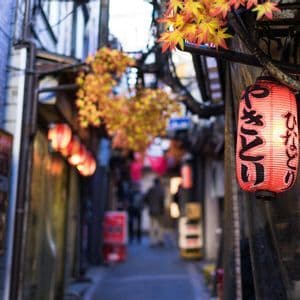 Gros plan sur une lanterne japonaise en papier rouge illuminée et calligraphiée, suspendue dans une ruelle étroite de ville décorée de feuilles d'automne.