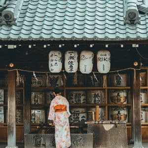 Eine Person in einem geblümten Kimono steht vor einem traditionellen japanischen Gebäude mit Papierlaternen und dekorativen Fässern.