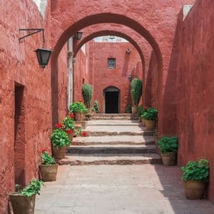 Un sendero de piedra con escaleras asciende a través de un estrecho corredor con paredes y arcos rojos, flanqueado por geranios en macetas.