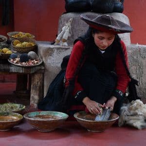 Une femme en tenue traditionnelle râpe une racine dans un bol pour fabriquer de la teinture naturelle, entourée de laine brute et de fils colorés.