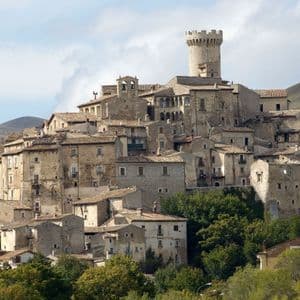 Un antico borgo in pietra con una torre rotonda costruito su una verde collina sotto un cielo parzialmente nuvoloso.