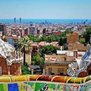Edifici fantasiosi, rivestiti a mosaico, che si affacciano su un vasto panorama urbano che incontra il mare sotto un cielo azzurro e limpido.