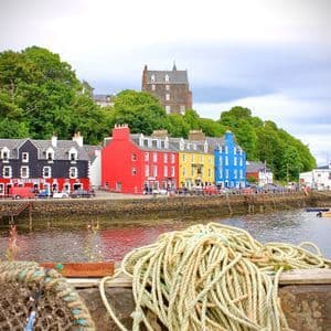 Une rangée de bâtiments aux couleurs vives longe le front de mer d'un port, avec d'épaisses cordes nautiques enroulées sur un quai au premier plan.