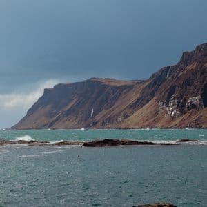 Une falaise brune et accidentée avec plusieurs cascades surplombe une mer turquoise agitée sous un ciel couvert.