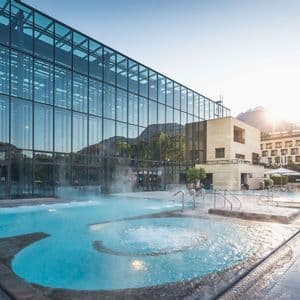 Piscina termale all'aperto con vapore che sale dall'acqua, accanto a un moderno edificio con facciata in vetro all'alba.