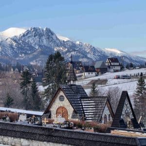 A stone chapel and village in a snowy valley at the foot of a large, snow-capped mountain range.