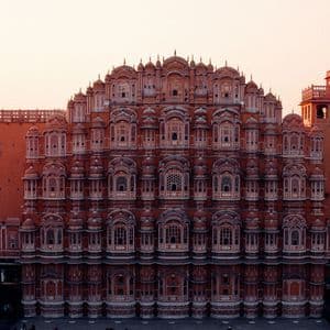 Die symmetrische Fassade eines prunkvollen, rötlichen Sandsteingebäudes mit Hunderten von kleinen Fenstern und Balkonen unter einem blassen Sonnenuntergangshimmel.
