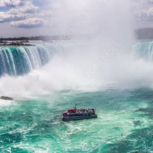 A tour boat sails on turbulent turquoise water near the base of a massive, horseshoe-shaped waterfall under a cloudy sky.
