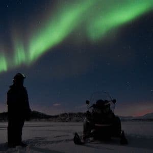 Una persona junto a una moto de nieve en un paisaje nevado nocturno, contemplando la aurora boreal verde que llena el cielo estrellado.