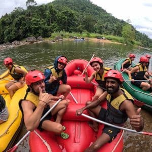 A WeRoad group trip poses for a photo in inflatable rafts on a river, wearing red helmets and life vests with a forested hill behind them.