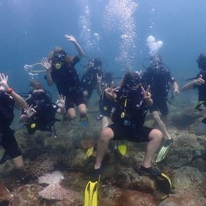 A WeRoad group trip in full scuba gear poses for an underwater photo, making hand signs above a rocky reef.