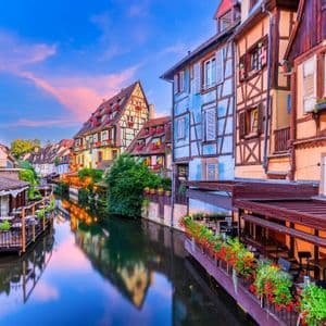 Edifici colorati a graticcio con balconi fioriti si affacciano su un canale tranquillo, riflettendosi nell'acqua al tramonto.