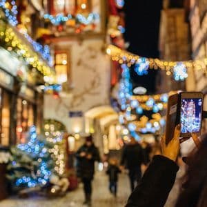 A person holds up a smartphone to photograph a narrow street decorated with colorful Christmas lights at night.