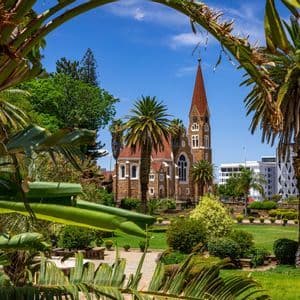 Una chiesa in pietra con una guglia rossa si erge in un parco curato incorniciato da palme tropicali sotto un cielo azzurro e limpido.