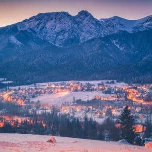 Un village enneigé aux lumières scintillantes niché dans une vallée au pied d'une grande chaîne de montagnes au crépuscule.