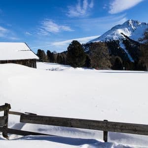 Eine rustikale Holzhütte und ein Zaun sind in einer tief verschneiten Berglandschaft unter strahlend blauem Himmel.